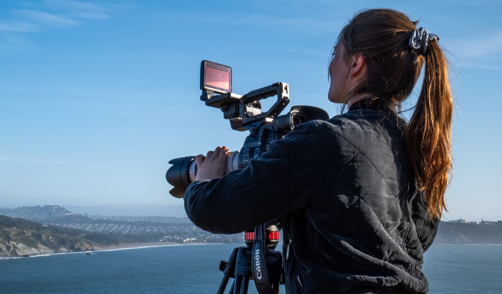 A photo of a woman with long hair tied back in a ponytail, with her back to the camera, looking into a video camera.