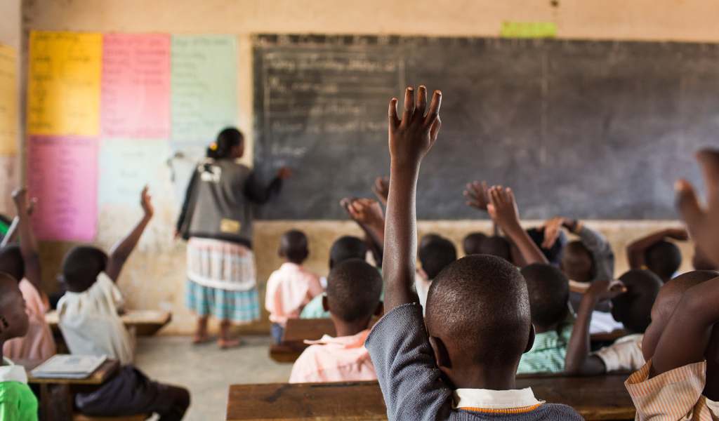 Children raise hands while sitting at wooden desks facing a large blackboard. A female teacher stands at the front of the class writing on the blackboard. 