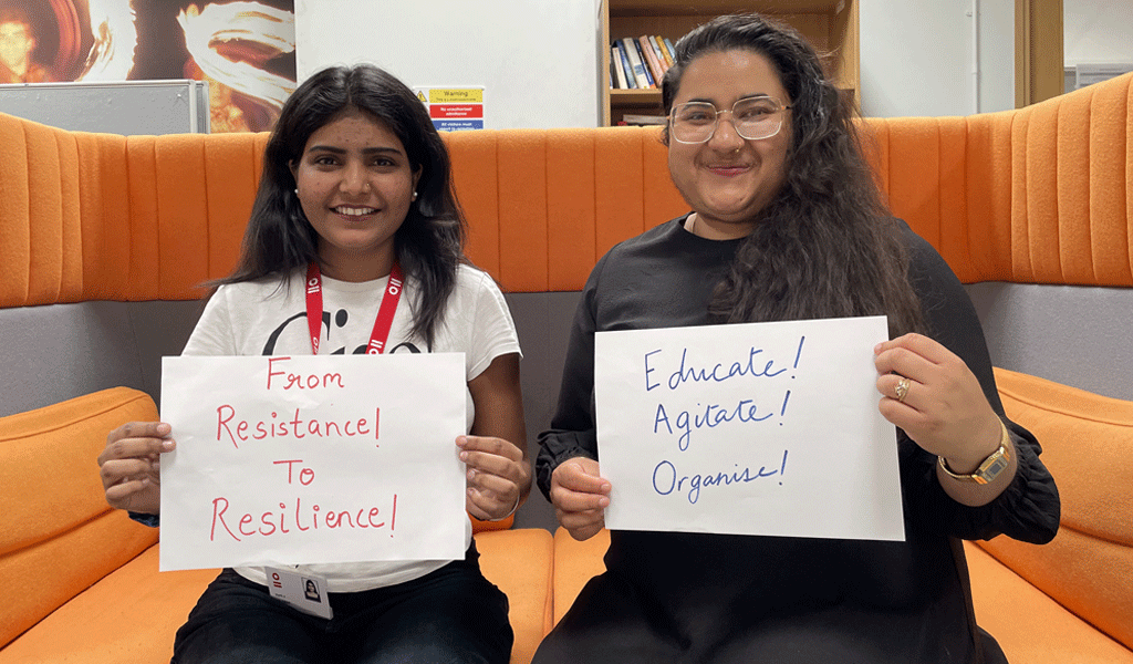 This is a photo of Aarti Rajput and Chandni Sai Ganesh sitting on a orange sofa. They are both holding signs with the words: From Resistance to Resilience and Education! Agitate! Organise!