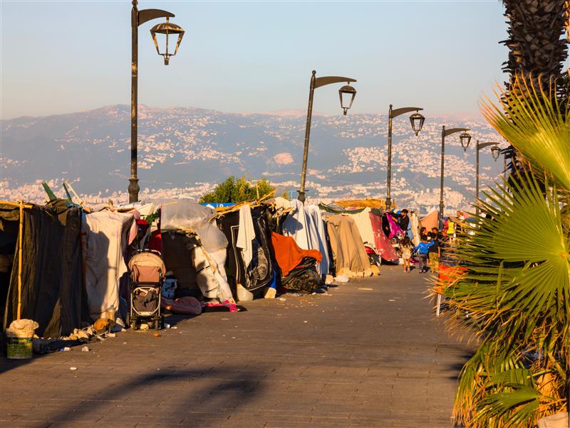 A photo of temporary housing in the form of shacks along a paved street in Beirut. The shacks are made of various materials. There are street lamps above the shacks and a large hill with more houses on it far in the distance.
