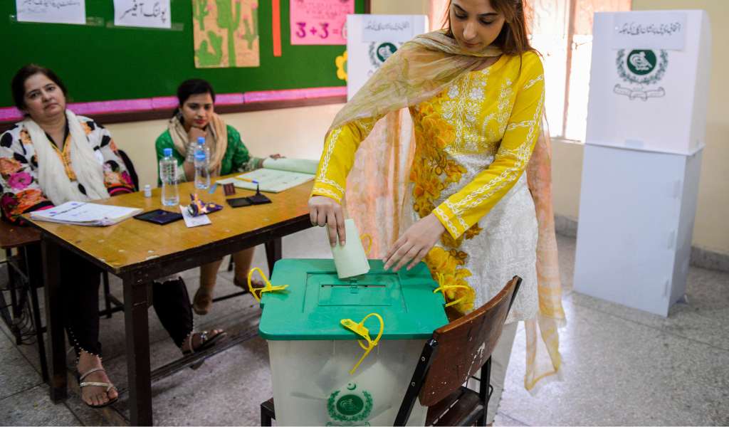 A woman in a yellow top places a white voting card into the top of a plastic voting box with a green lid. Two women sit at a desk in the background. 