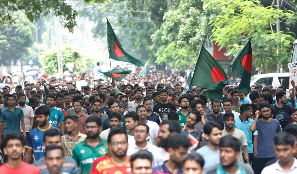 A dense crowd of people marching outdoors. 3 green flags with red circles are being waved. Trees are in the background and the crowd stretches back to the horizon.