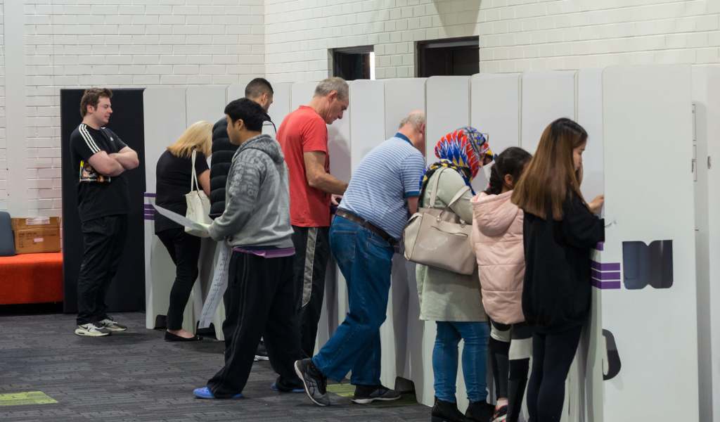A row of people who are each standing at a white cardboard booth. Their backs are to the camera. The room they are in has white walls and grey carpet.