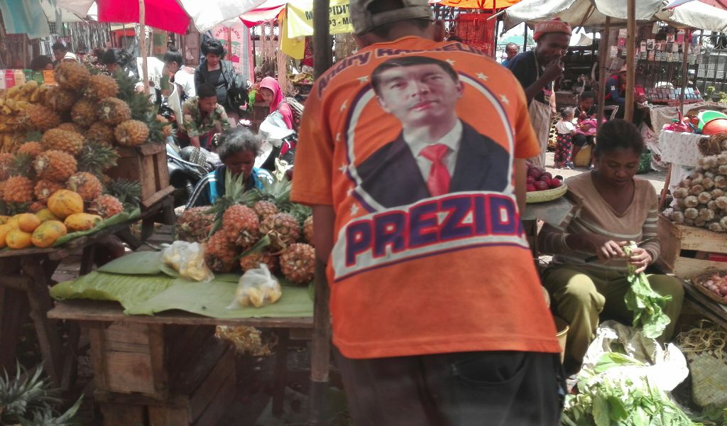The back of a man wearing an orange t.shirt with a big photo of the President Rajoelina, with the word 'Prezid' showing. He is facing a fruit stall in a market.