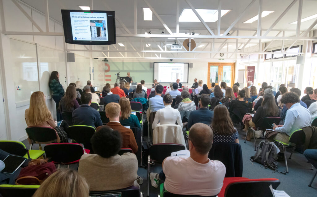 Audience seated in a conference room at IDS listening to a panel presentation during the BASIC Research International Conference, with slides displayed on large screens.