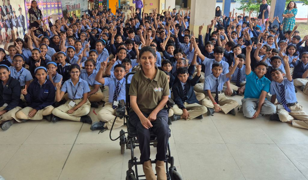 A young Indian woman sits in a wheel chair smiling at the camera. Many rows of school children are in the background wearing blue shirts.