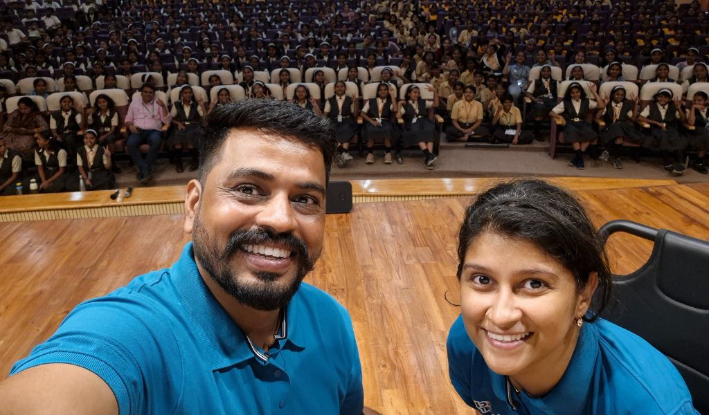 A young woman and man wearing blue shirts smiling at the camera, with rows of school children in the background.