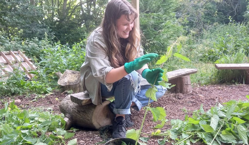 A young woman in a green top wearing green gloves, sitting on a wooden log and holding a plant