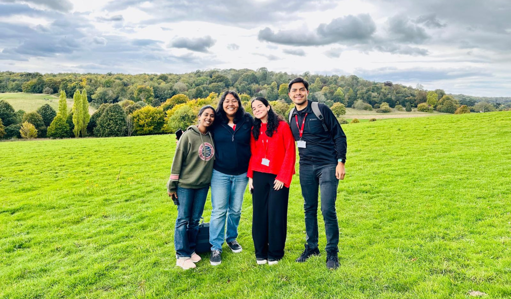 Four people stand next to each other smiling. They are standing outside on green grass and behind them is a hill with trees and the sky has clouds with the sun shining behind them.