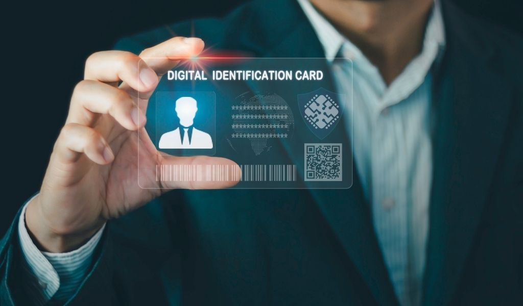 A man in a suit against a dark background holds up a digital impression of an ID card. The card says Digital Identification Card with a head and shoulder image and a QR code. 