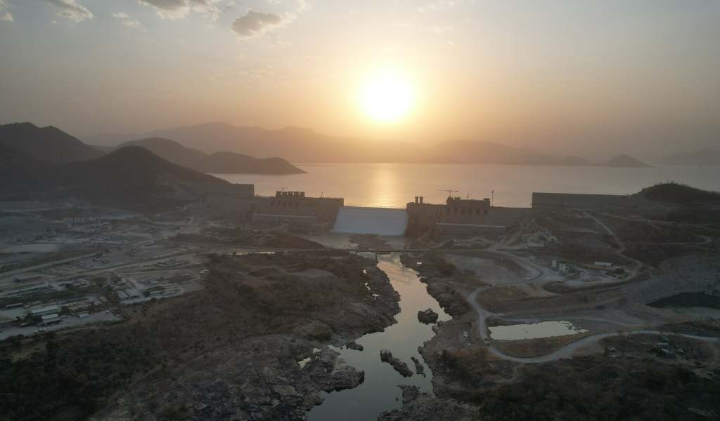 Sunset above a body of water with a dam in the middle ground and a small river flowing from it into the foreground with greenery and buildings either side of the river bank.