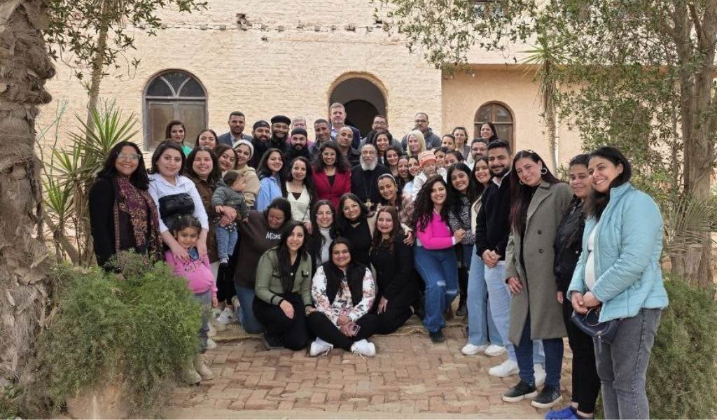 About 50 people in a group photo outdoors. Most are standing, some at the front are sitting. A stone building is in the background and trees can be seen at the sides.