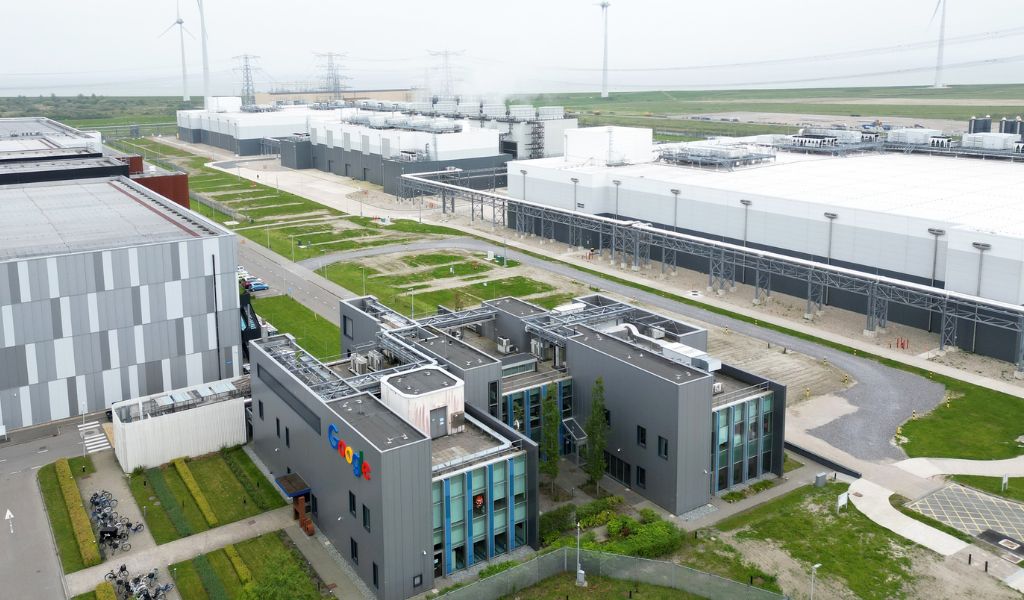 An aerial photo of large grey and white industrial buildings. 'Google' is just visible on the side. The buildings are surrounded by squares of green grass. 