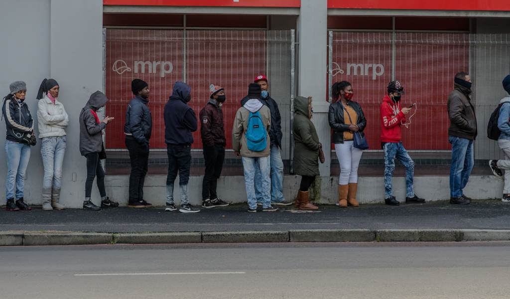 People lined up in single file on a pavement in front of a shop window. Some are wearing face masks.
