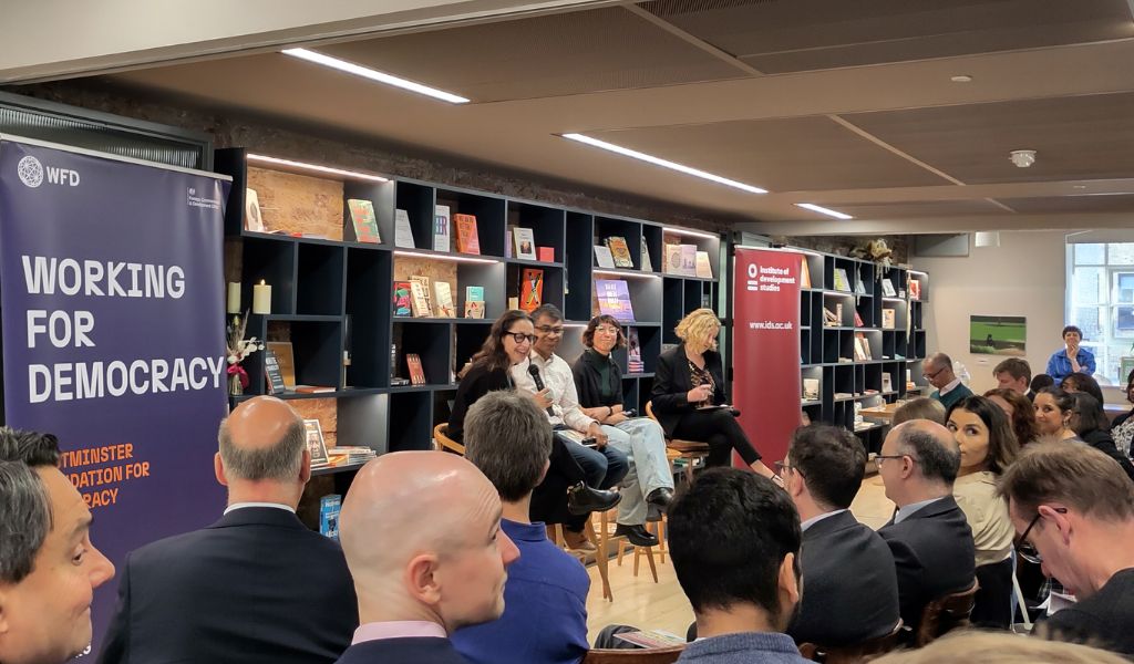 Four people on stools at the front of a crowded room. Book shelves are behind them and banners either side. The nearest says 'Working for democracy'.