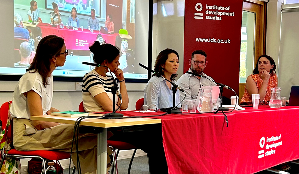 A panel discussion taking place at the Institute of Development Studies (IDS), with five speakers seated behind tables covered with red IDS-branded tablecloths. Microphones, water jugs and glasses are arranged in front of the speakers. A projection screen behind the panel displays a live view of the discussion, and an IDS banner featuring the organisation’s logo and website stands alongside the panel. The event is being held in a bright conference room with attendees visible in the audience.