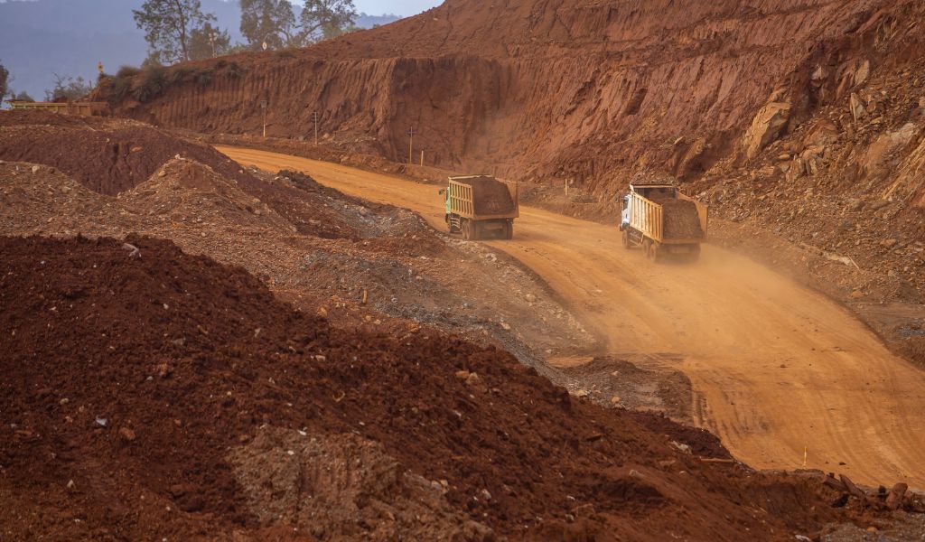 Two lorries driving through a sandy track between red earth mounds, leaving blooms of dust behind them. 