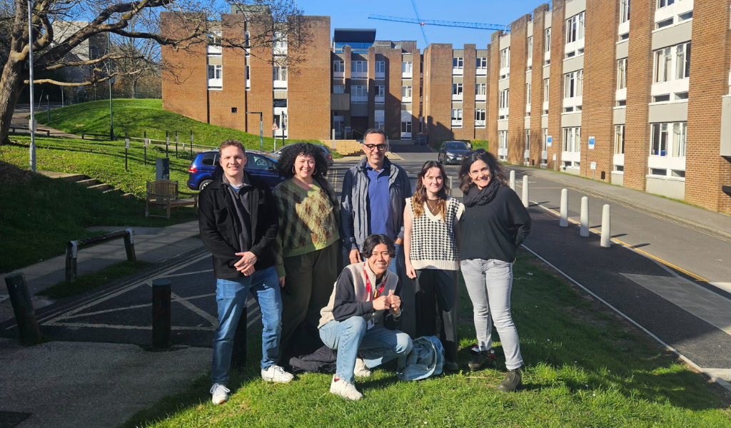 Six people stand together on a grassy area outside campus accommodation buildings on a sunny day. One person is crouching at the front while five others stand behind. Brick residence blocks, a parked car, pathways, trees and a clear blue sky are visible in the background.