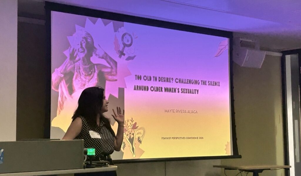 A presenter stands beside a projected conference slide titled “Too Old to Desire? Challenging the Silence Around Older Women’s Sexuality” by Mayte Rivera Alaga at the Feminist Perspectives Conference 2026. The presenter gestures towards the screen while speaking in a lecture room with desks and presentation equipment visible in the foreground.