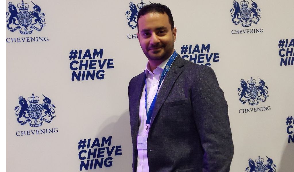 A medium shot of a man with short, dark curly hair and a slight beard, smiling at the camera. He is wearing a grey textured blazer over an unbuttoned white shirt, and a blue lanyard with an ID badge is around his neck. He is standing against a white backdrop patterned with the dark blue royal coat of arms of the United Kingdom and the word "CHEVENING" below it, alongside the hashtag "#IAMCHEVENING" in a bold sans-serif font.