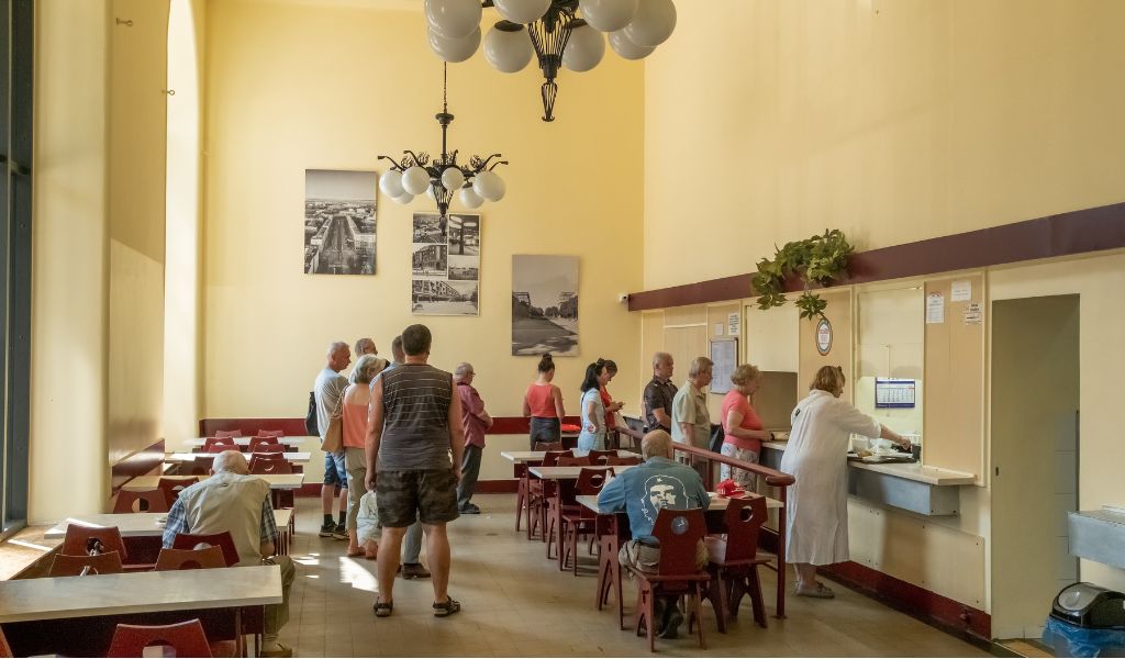 A canteen with large ceiling and pale yellow walls. A queue of people wait for food served out of windows in the wall. Tables in rows with red plastic chairs.