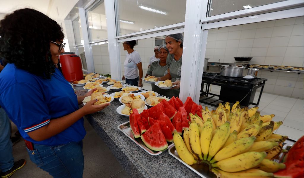 trays if brightly coloured yellow bananas and red watermelon on a counter. A young woman is standing in front of it. Three women wearing hairnets are on the other side serving. 