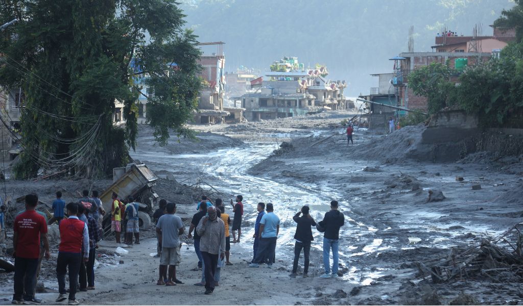 A group of men stand looking down at the remains of flood water curving down the remains of a town. Severely damaged buildings can be seen in the distance.
