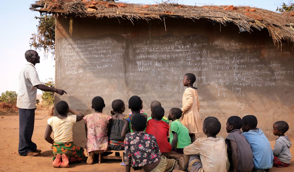 Around 15 children sit on a sandy floor, outside, facing the side of a small building, with a grass roof, being used as a chalk board. It has writing of words on it. An adult teacher stands to the side.